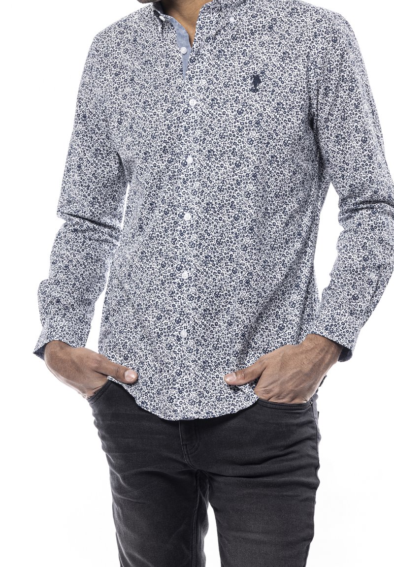 Homme portant une chemise blanche à boutons avec un motif floral bleu et un jean noir, les mains dans les poches, sur un fond blanc.