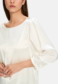 White, silky blouse with a round neckline, puffed sleeves, and elastic cuffs. Smooth texture and subtle sheen enhance its elegant appearance.