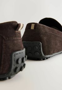 Barna velúr slip-on cipők gumitalppal. Jellemzőik között szerepel egy texturált fekete sarok és egy minimális csíkozás a sarkon.