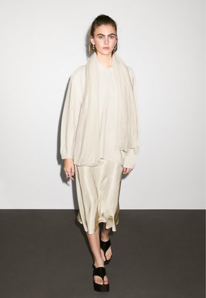 Beżowy, oversize sweter, kremowa satynowa spódnica z falistym dołem i czarne sandały na platformie. Lekki szalik zarzucony na szyję.