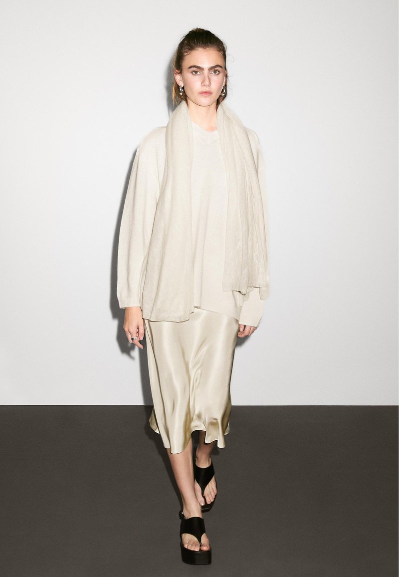 Beige oversized sweater, cremet satin slip nederdel med bølget kant og sorte platform sandaler. Letvægts tørklæde hængende om halsen.