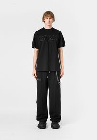 Camiseta negra de gran tamaño con logotipo en relieve, combinada con pantalones negros con bolsillos con cremallera y acentos de cadena, usados con calzado negro texturizado.