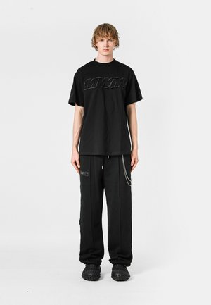 Camiseta negra de gran tamaño con logotipo en relieve, combinada con pantalones negros con bolsillos con cremallera y acentos de cadena, usados con calzado negro texturizado.
