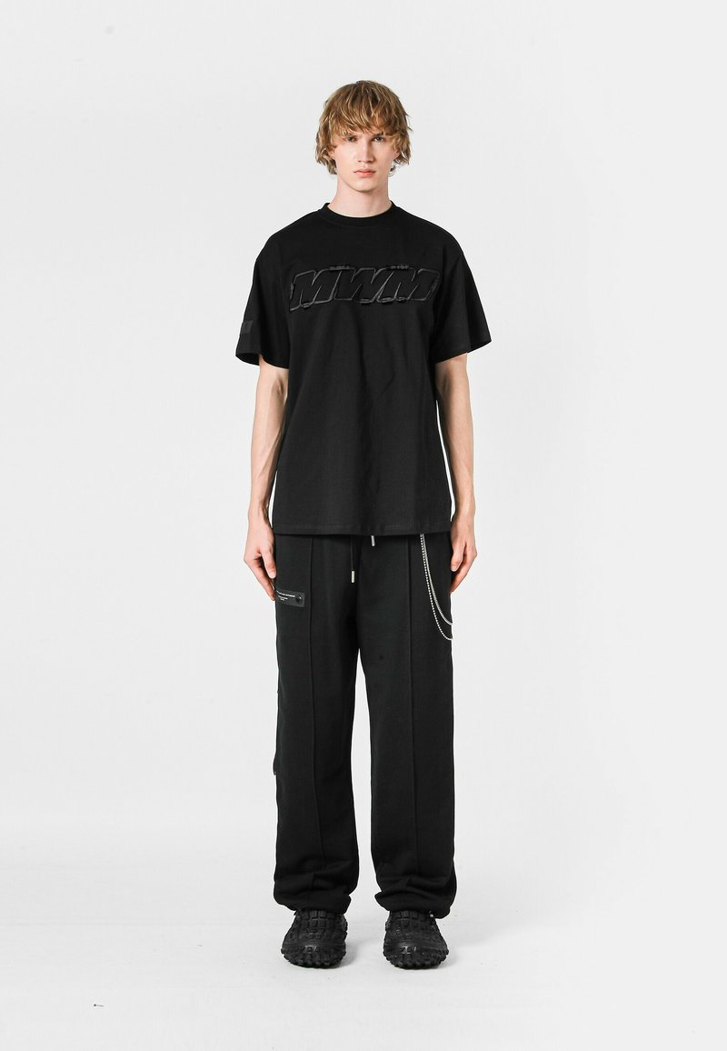 Camiseta negra de gran tamaño con logotipo en relieve, combinada con pantalones negros con bolsillos con cremallera y acentos de cadena, usados con calzado negro texturizado.