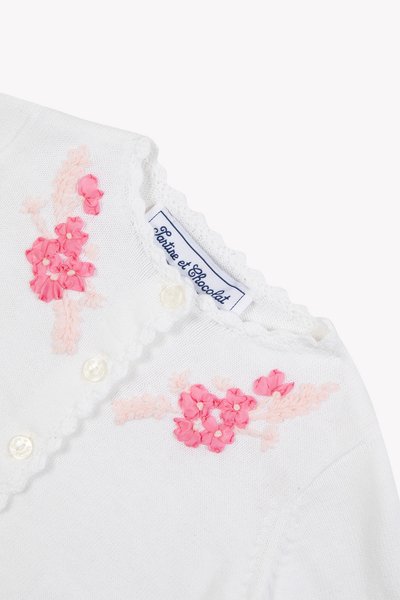 Λευκή πλεκτή ζακέτα με ροζ κεντήματα λουλουδιών, με στρογγυλά κουμπιά και λαιμόκοψη με κυματιστά σχέδια. Η ετικέτα γράφει "Partie et Chocolat."