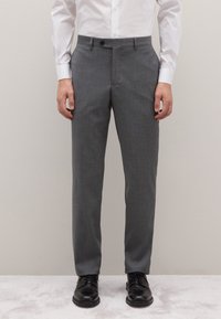 Pantaloni grigi su misura in tessuto leggero, con frontale piatto, passanti per cintura e chiusura con un bottone singolo. Abbinati a scarpe nere.