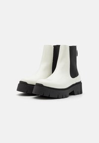 Cizme ankle albe și negre cu o talpă groasă neagră, textură netedă, panouri laterale elastice și o bucată de prindere în spate.