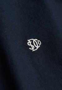 Close-up of dark blue fabric with small white embroidered logo featuring stylized letters "SD" or "ED" centered.