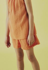 Completo in tessuto arancione caratterizzato da una canotta senza maniche e pantaloni a pantaloncino. Il tessuto ha un aspetto sgualcito, con un design a strati.