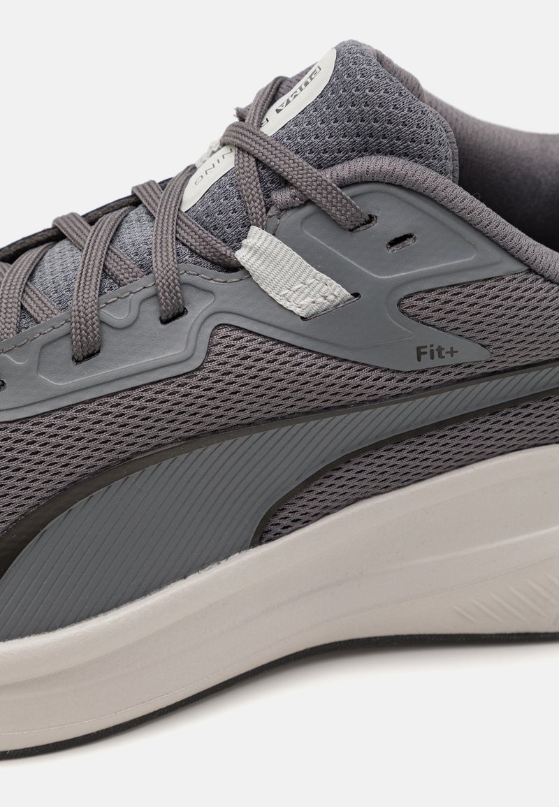 Primer plano de una zapatilla deportiva gris con tela de malla, suela blanca gruesa, etiqueta "Fit+" y cordones atados sobre la lengüeta acolchada.