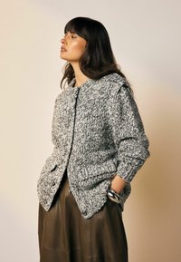 Grauer und weißer gestrickter Cardigan mit langen Ärmeln, runder Halslinie und zwei vorderen Taschen, kombiniert mit einem braunen Lederrock.
