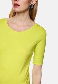 Helles Gelb, tailliertes Oberteil mit weitem Ausschnitt und Dreiviertelärmeln. Der Stoff wirkt glatt mit einem leichten Glanz. Silbern, rechteckige Ohrringe.