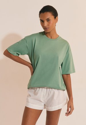 Donna in piedi con una mano sui fianchi, indossa una maglietta verde a maniche corte e larga e pantaloncini bianchi, su uno sfondo beige semplice.