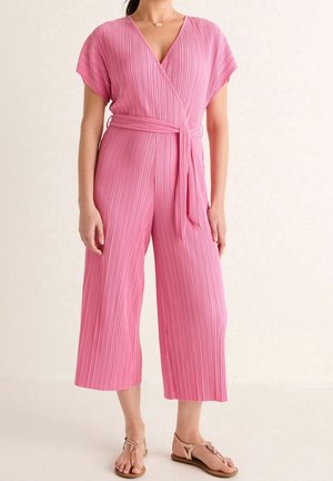 Kobieta w różowym plisowanym kombinezonie z szerokimi nogawkami, krótkimi rękawami i paskiem do wiązania w talii, w połączeniu z beżowymi sandałami.