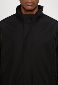 Černá bunda na zip s vysokým límcem, vyrobená z hladkého, lehkého materiálu s jemným šitím a bez viditelných kovových prvků.