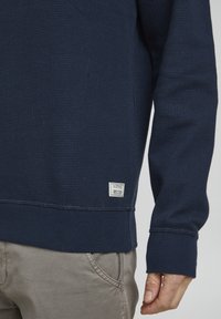 Tumši zila teksturēta džemperis ar ribinātām piedurknēm un apakšmalu. Uz apakšējā kreisā stūra ir neliels marķējums ar zīmola un projekta informāciju.