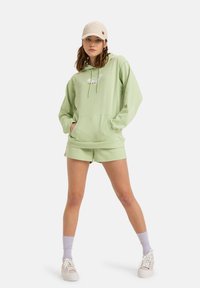 Felpa verde chiaro con tasca frontale e logo, abbinata a pantaloni corti coordinati. Include un cappello beige, scarpe da ginnastica bianche e calzettoni lilla.