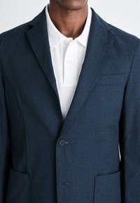 Sacou texturat de culoare albastru închis, cu o croială ajustată, închidere cu un singur nasture și un buzunar; stilizat cu un tricou polo alb dedesubt.