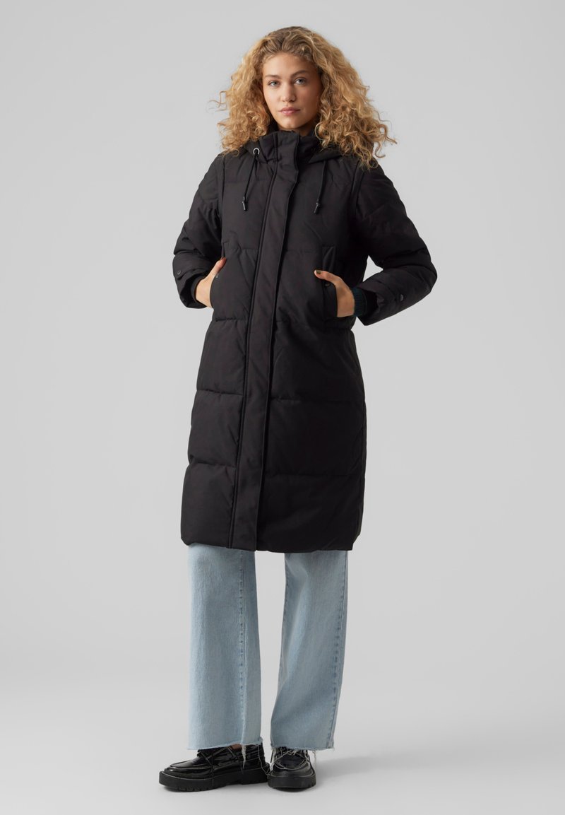 Svart pufferjakke med hette, som har glidelås foran, to sidelommer og quiltet design. Kombinert med lyseblå vide jeans og svarte sko.