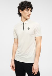 Ανδρικό μπεζ πόλο μπλουζάκι με φερμουάρ στο γιακά, λογότυπο στο αριστερό στήθος, κοντά μανίκια και λείο ύφασμα, σε συνδυασμό με σκούρο παντελόνι.