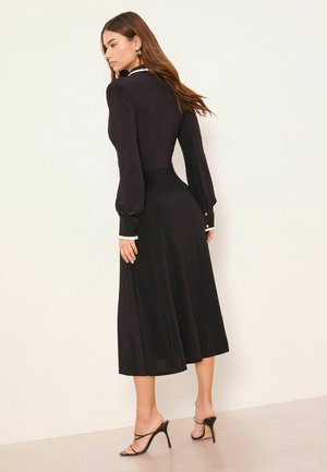 Schwarzes langärmeliges Kleid mit tailliertem Oberteil, ausgestelltem Rock, weißem Kragen und goldfarbenen Knopfakzenten an den Manschetten. Glatte Stofftextur.