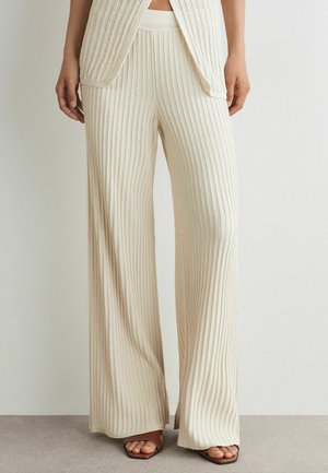 Kobieta ubrana w kremowe, prążkowane, szerokie spodnie dzianinowe oraz pasujący bezrękawnik, stoi ze swobodnie opuszczonymi rękami.