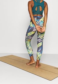 Teal sportbh med urtag på ryggen, som matchar livfulla leggings med tropiskt bladmönster i mörka toner på en yogamatta.