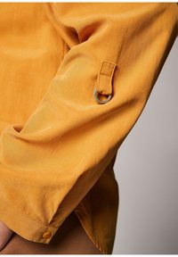 Camicia gialla a maniche lunghe con una texture liscia, caratterizzata da un dettaglio in metallo argentato a forma di anello e pulsante sul polsino.