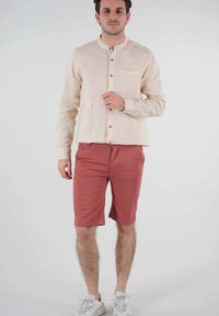 Chemise à manches longues beige clair avec fermeture à boutons et poche poitrine, associée à un short rose. Le mannequin porte des baskets blanches avec des lacets.