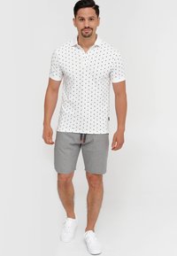 Hvid polo t-shirt med sort prikket mønster, korte ærmer og krave, parret med grå stribede shorts og hvide sneakers.