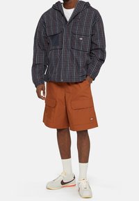 Man som bär en marinblå rutad jacka med huva, rostfärgade cargo-shorts, vita strumpor och vita Nike-sneakers med brun swoosh.