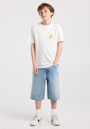 Junge steht mit einer Hand in der Tasche, trägt ein weißes T-Shirt mit gelbem Logo, kniehohe Jeansshorts, weiße Socken und Turnschuhe.