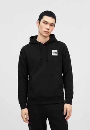 Junger Mann trägt einen schwarzen The North Face Hoodie mit Fronttasche und Logo auf der Brust, steht vor einem schlichten hellen Hintergrund.