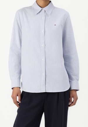 Jasnoniebieska i biała cienko prążkowana koszula na długi rękaw z guzikami i kołnierzykiem, noszona z czarnymi spodniami, bez widocznych dodatków.