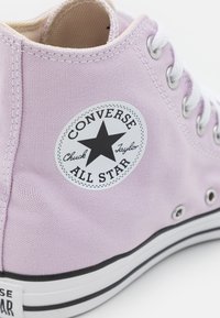 Sneaker de cano alto em tela lilás claro com atacadores e ilhós brancos, apresentando um remendo com o logo de estrela preta rotulado "Converse All Star".