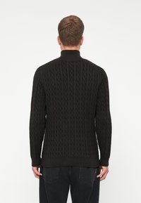 Dunkelbrauner Strickpullover mit strukturiertem Zopfmuster, hohem Kragen und gerippten Bündchen und Saum, von hinten gezeigt.