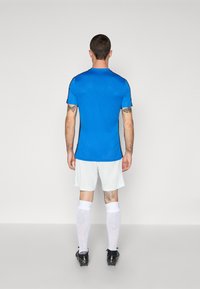 Modré sportovní tričko s krátkým rukávem a černými akcenty, bílé šortky, bílé ponožky po kolena a černé kopačky. Tetování viditelné na pažích.