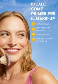 Primer leggero in una formula trasparente. Modello sorridente, che tiene un pennello per il trucco indossando abbigliamento rosa pastello e collane a strati contro uno sfondo di cielo blu.