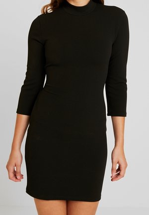 Frau trägt ein eng anliegendes, schwarzes, knielanges Kleid mit dreiviertel Ärmeln und einem hohen Ausschnitt, steht vor einem einfarbigen Hintergrund.