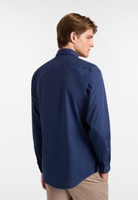 Mornarska srajca z dolgimi rokavi, razširjenim ovratnikom, gladko strukturo in gumbi na manšetah. Nazaj je vidna rahlo podaljšana oblika in prilegajoča zasnova.