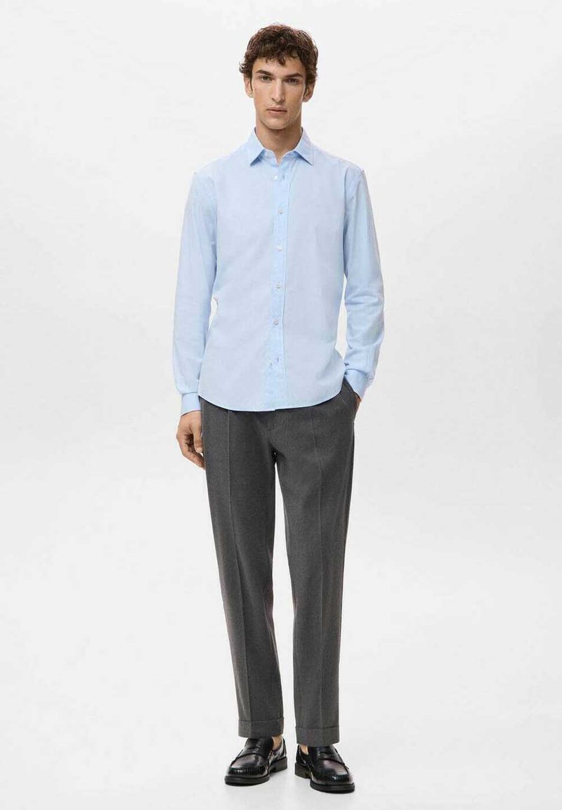 Camicia azzurro chiaro a maniche lunghe con colletto abbottonato, abbinata a pantaloni grigi su misura e scarpe nere, su sfondo bianco.