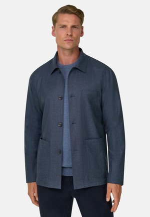 Homme portant une veste boutonnée gris foncé par-dessus un pull bleu, debout devant un fond clair uni, face à l'avant.