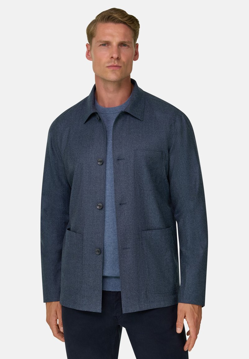Homme portant une veste boutonnée gris foncé par-dessus un pull bleu, debout devant un fond clair uni, face à l'avant.