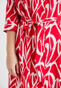 Φόρεμα σε κόκκινο και λευκό μοτίβο με ζώνη, κοντά μανίκια και κουμπιά μπροστά, κατασκευασμένο από ελαφρύ ύφασμα με λεία υφή.