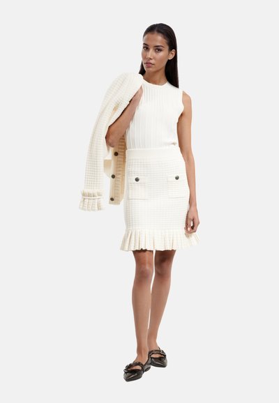 Top blanc sans manches en tricot avec des côtes verticales ; jupe crayon assortie avec une texture en damier et un ourlet froncé ; ballerines noires ; cardigan léger drapé sur le bras.