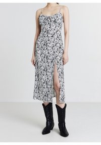 Robe midi à motifs noir et blanc avec une fente latérale, des bretelles fines et un détail noué à l'avant, portée avec des bottes de cowboy noires.