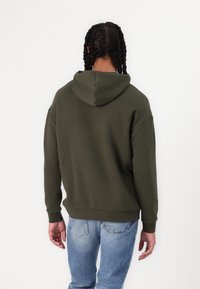 Olive green hooded sweatshirt with a relaxed fit, ribbed cuffs, and hem. Textured fabric with a smooth finish. Worn with light blue jeans.