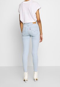 Osoba v světle modrých skinny džínách, bílem tričku s krátkým rukávem a bílých kotníkových botách, stojící na jednobarevném světlém pozadí.