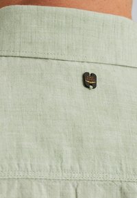 Lichtgroene shirtstof met een gestructureerde afwerking, voorzien van een zwart metalen label met de tekst "PME Legend" bevestigd aan de kraag.