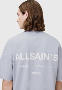 Tricou grafic de culoare gri deschis, fabricat din bumbac, cu un text mare alb pe spate care citește "ALLSAINTS" și "LONDON" într-un font îndrăzneț.